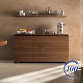 Credenza moderna piccola a tre porte in legno di noce, collezione Modigliani di Piombini, con intaglio formella centrale. Ambientata in uno spazio cucina raffinato, con macchina del caffè e accessori da colazione. Logo centenario 1925–2025 in basso a destra.